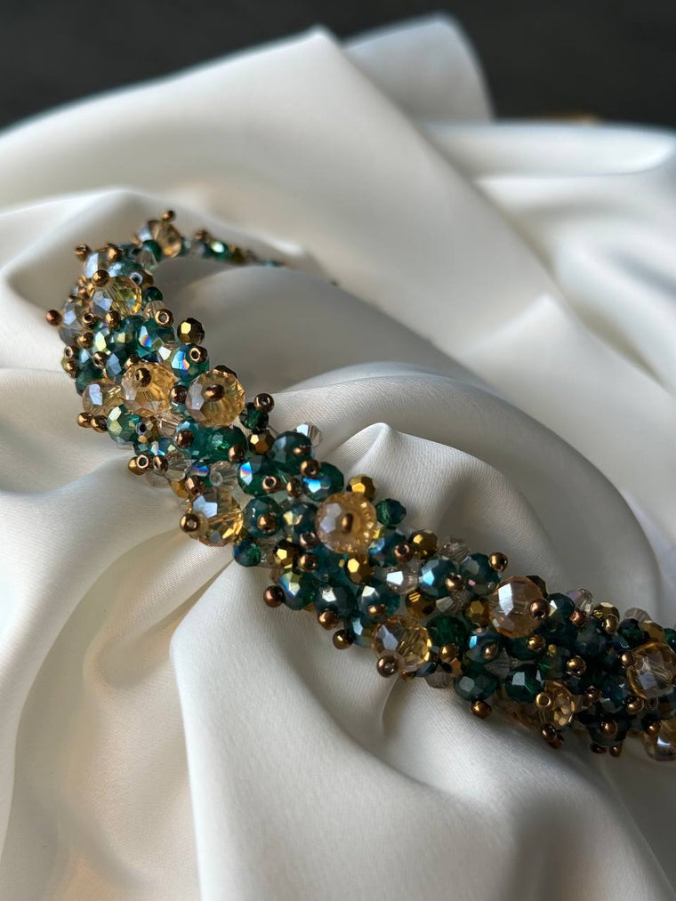 A close-up of a beaded headband featuring dark teal, gold, and brown faceted beads, resting on draped white satin fabric 