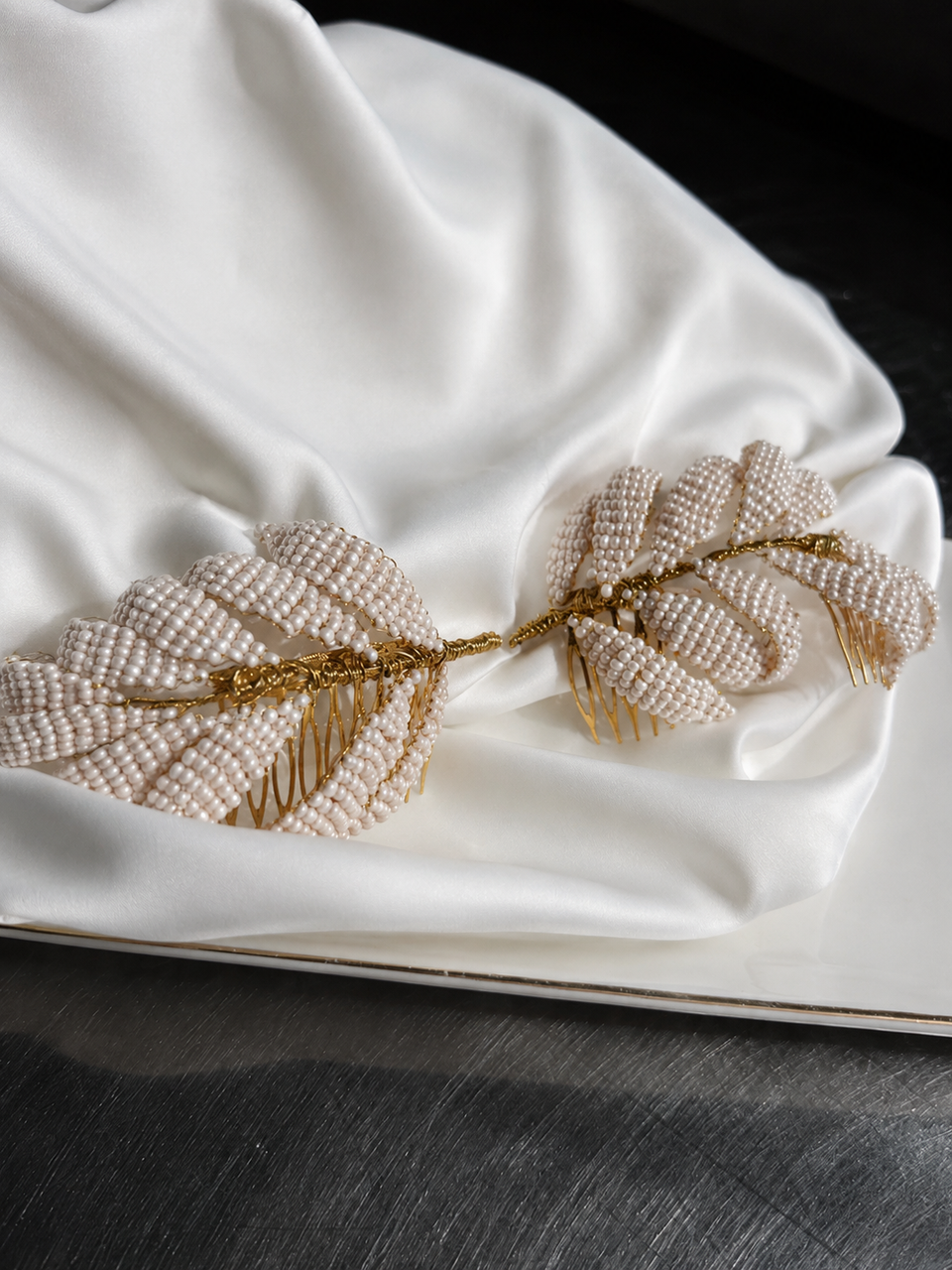 A beaded hair comb styled like two leaves connected by a gold wire branch, decorated with cream-colored pearls and gold metal accents, resting on draped white satin fabric.