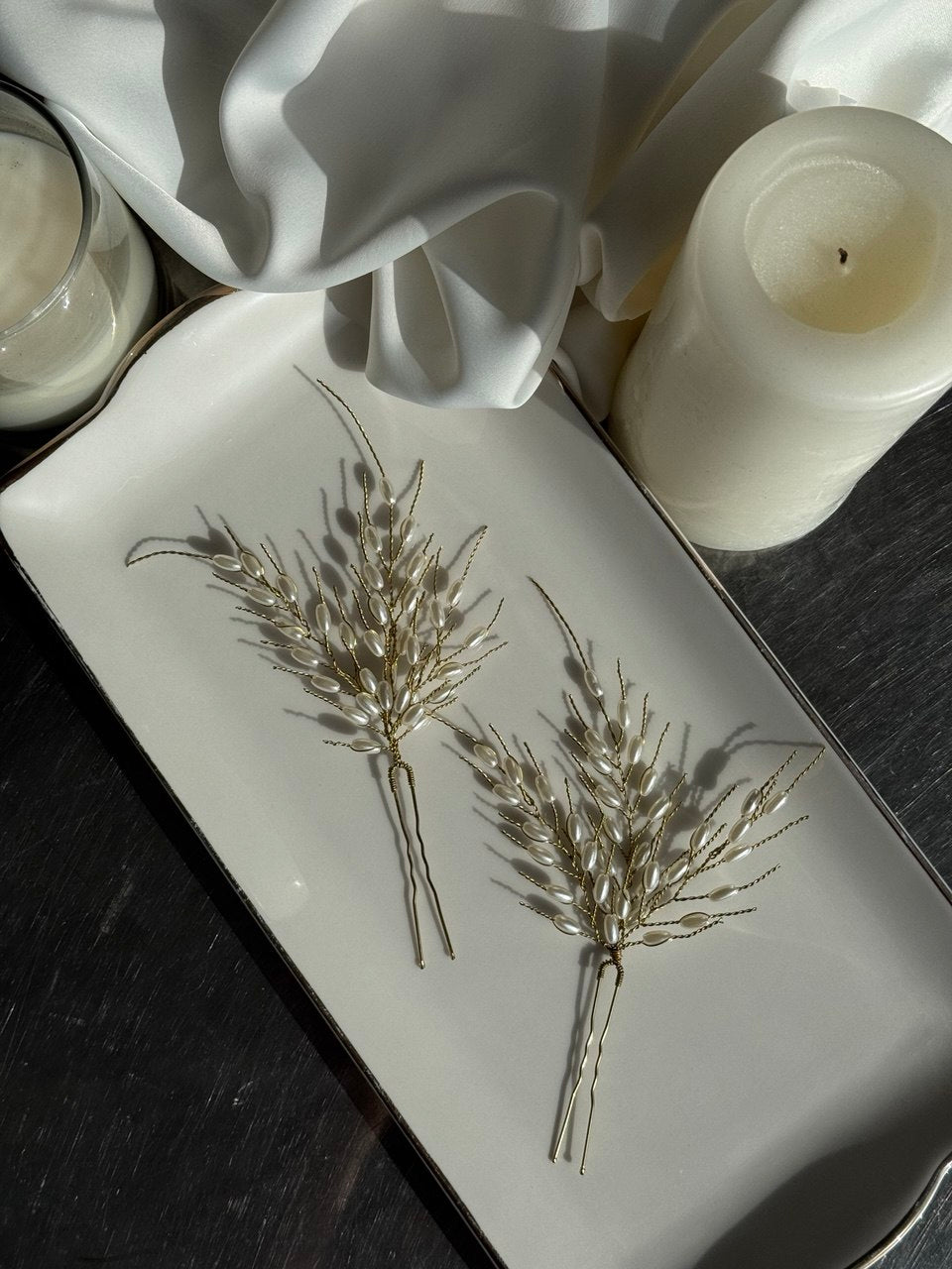 Two metallic wheat-like hair pins displayed on a white rectangular tray, surrounded by draped white fabric, a white candle, and a dark tabletop.