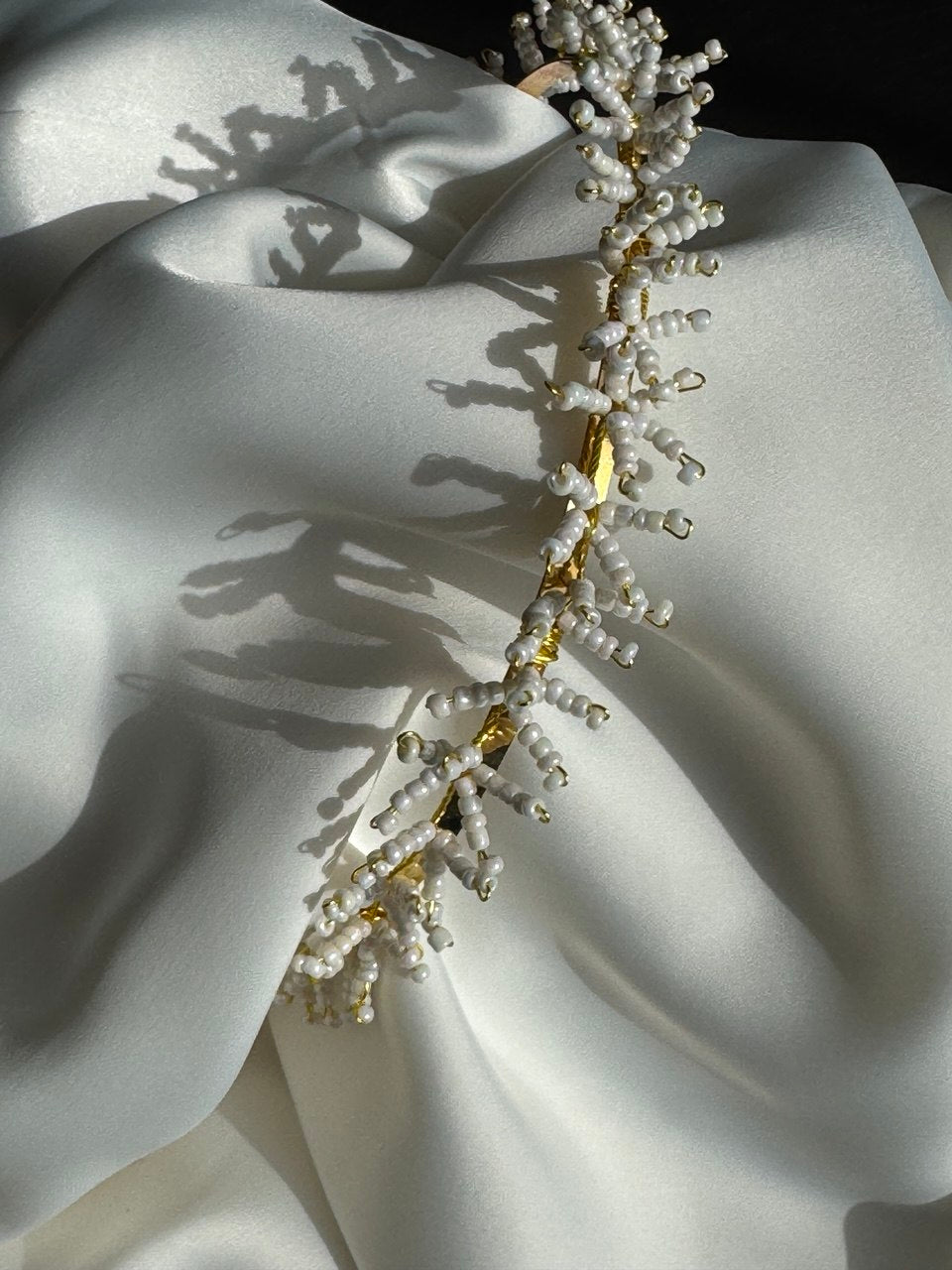 A beaded bridal headpiece with white pearl-like beads and gold-toned metal details, draped diagonally across draped white satin fabric with strong sunlight shadows.